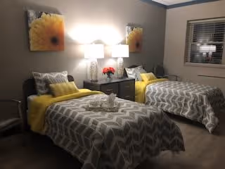 A cozy bedroom with two twin beds featuring gray and white chevron-patterned bedding and yellow accent pillows. A nightstand with two lamps and a vase of red flowers is positioned between the beds. Two framed pictures of yellow flowers hang on the wall above the nightstand. A window with blinds is visible on the right side.