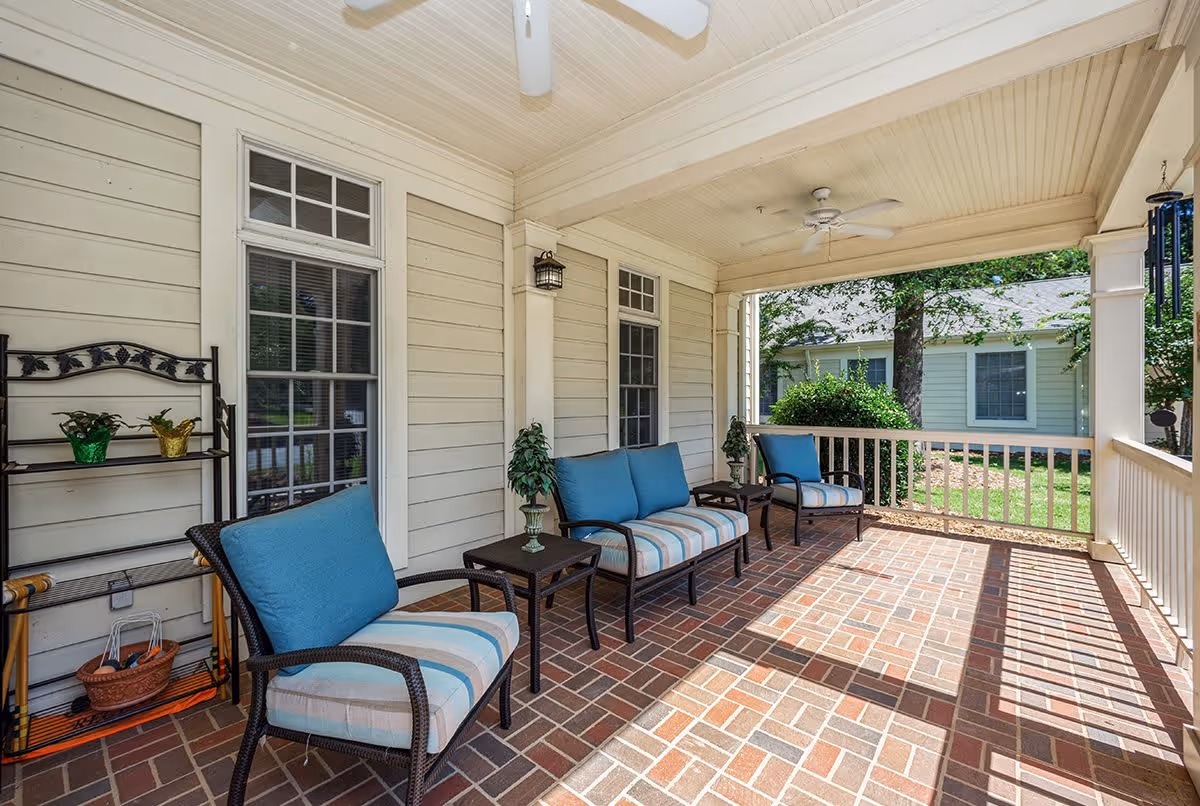 A covered outdoor porch area with brick flooring, white wooden railings, and ceiling fans. The porch is furnished with three cushioned chairs and two small tables, each table holding a small potted plant. There is a metal shelf with decorative items and plants against the wall. The porch overlooks a green yard with trees and a neighboring building.