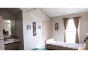 A small senior living bedroom with a single bed, a window with curtains, wall art, and an open sink/vanity area.