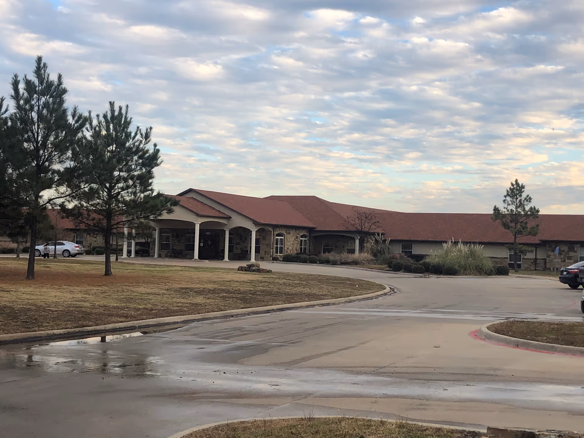 One-story senior living building with a covered porte-cochère entrance, nearby trees, a curved driveway and a cloudy sky.