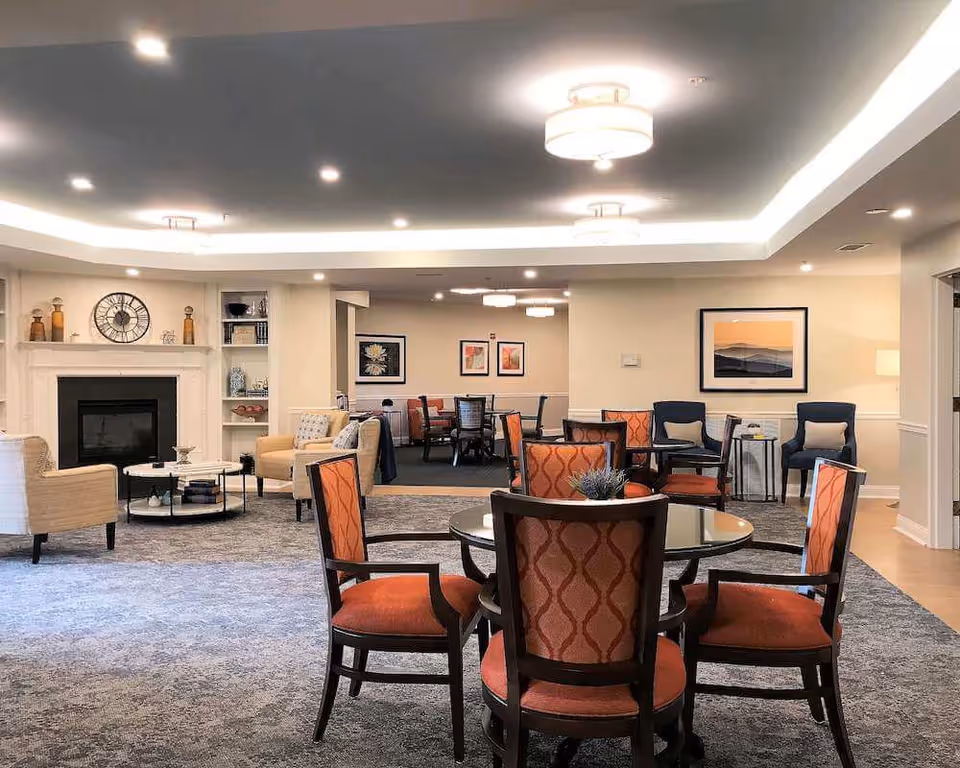 A spacious and well-lit common area in a senior living facility featuring a round table with four orange cushioned chairs in the foreground. In the background, there are additional seating areas with beige armchairs near a fireplace, blue chairs against the wall, and more tables and chairs further back. The room has a modern ceiling with recessed lighting and decorative light fixtures, along with framed artwork on the walls.