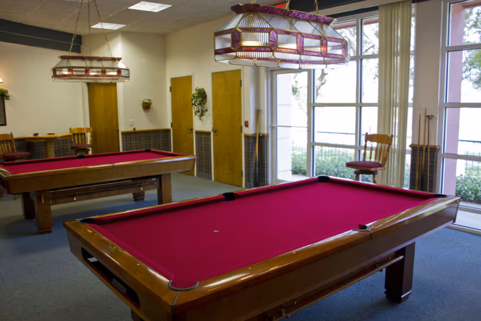 Interior room with two red felt pool tables, wooden chairs, and large windows letting in natural light. The room has hanging stained glass light fixtures above the pool tables and wooden doors along the wall.