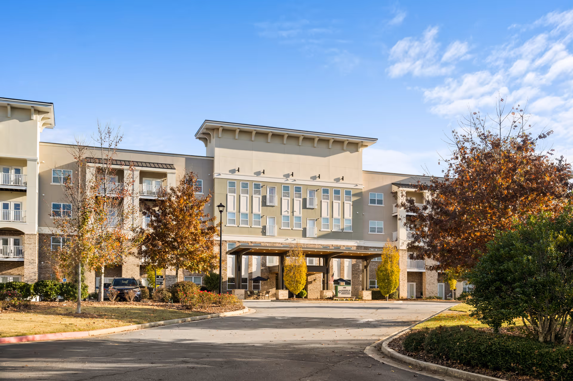 Exterior view of The Mansions at Alpharetta Senior Independent Living building with a covered entrance, surrounded by trees with autumn foliage and a clear blue sky.