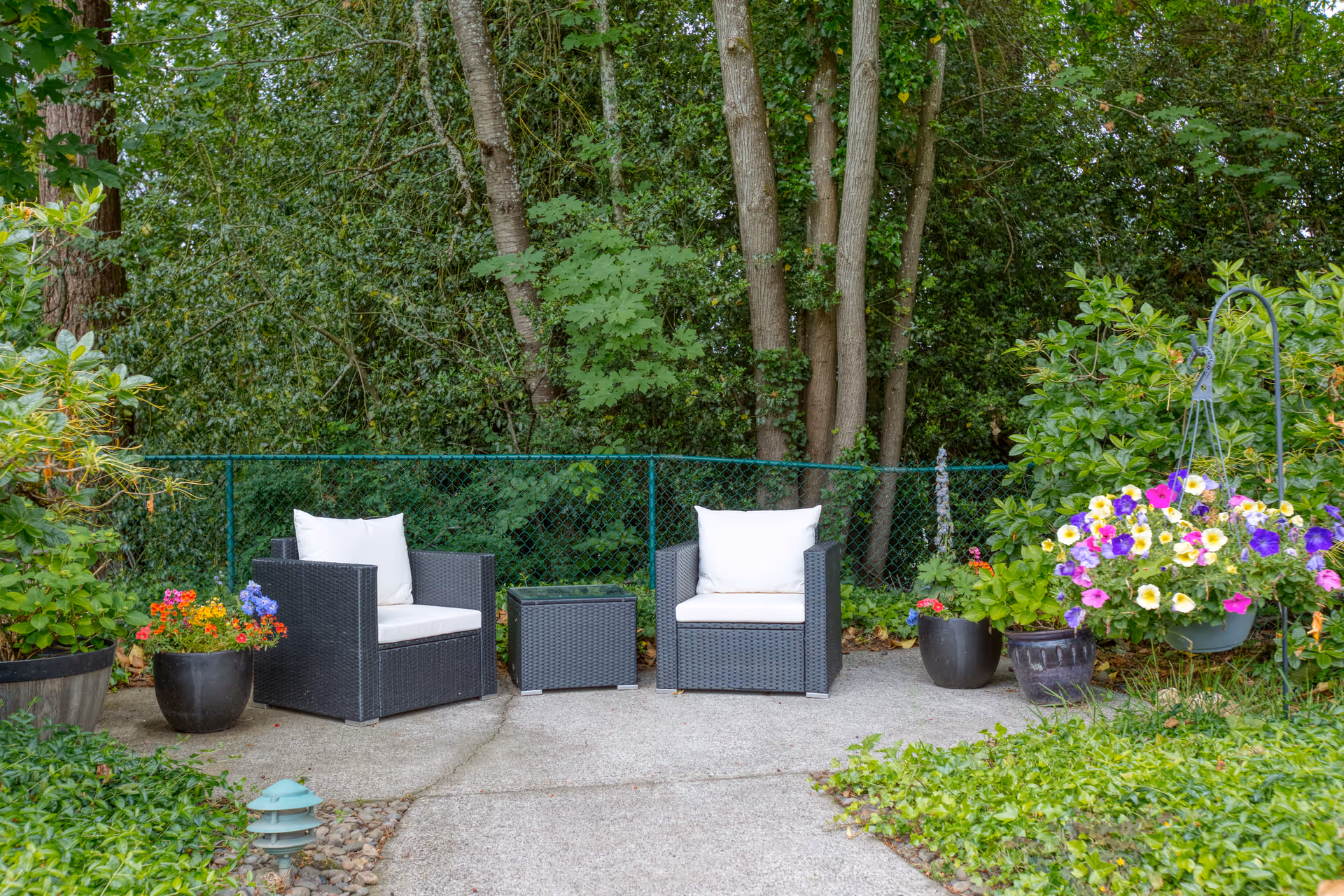 Two wicker patio chairs with white cushions and a small table on a concrete patio surrounded by potted flowers and trees.