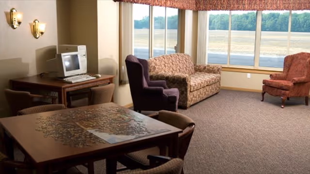 Sunlit common room with sofas and armchairs, a table with a partially completed puzzle, and a computer station by large windows.