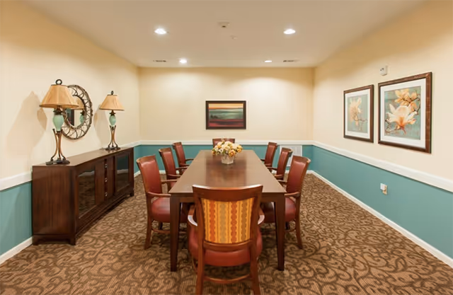 A long wooden table surrounded by upholstered chairs in a well-lit dining/meeting room with framed artwork and decorative lamps.
