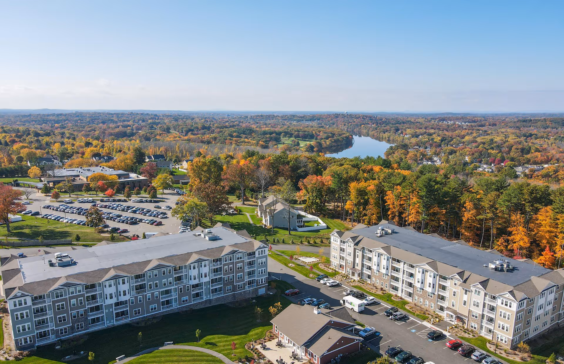 Aerial view of Riverside Woods by Pulte Homes senior living facility with multiple large residential buildings, parking lots, and surrounding trees showing autumn colors. A river is visible in the background under a clear blue sky.