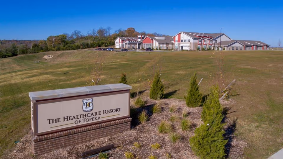 Wide exterior view of The Healthcare Resort of Topeka building situated on a grassy hill with a clear blue sky. In the foreground, there is a large sign with the facility's name surrounded by small shrubs and landscaping.