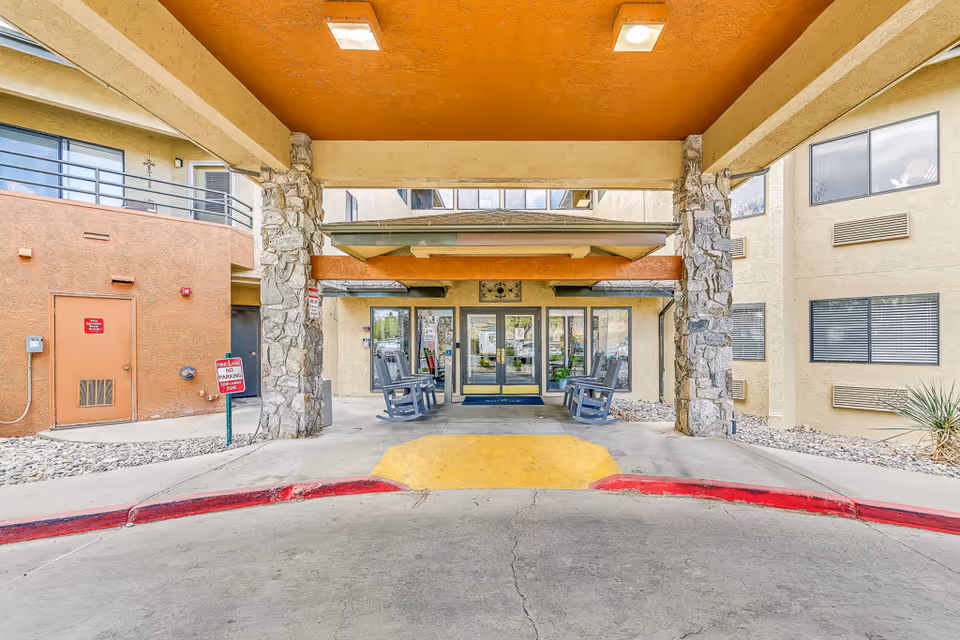 Covered entrance/porte-cochère with stone columns, rocking chairs and glass doors leading into the senior living building.