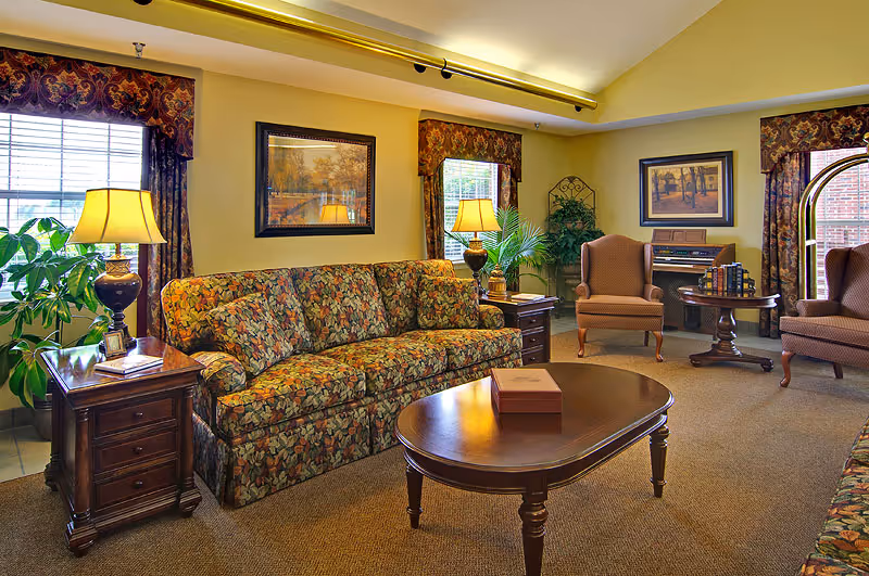 A cozy living room with floral patterned sofas, two armchairs, a wooden coffee table, side tables with lamps, framed artwork on the walls, and large windows with decorative curtains letting in natural light. There are also some green plants and a small piano with books on it in the corner.