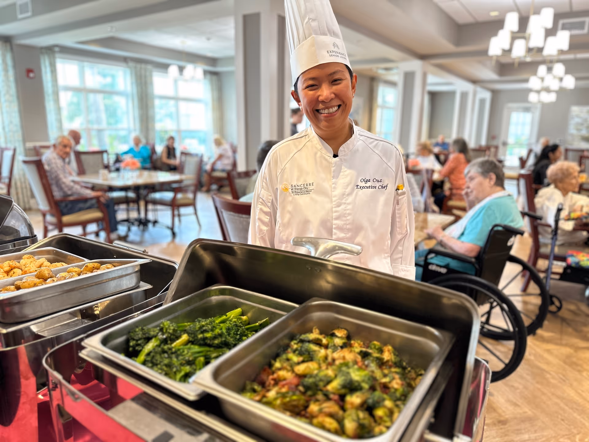 A smiling chef wearing a white chef's coat and hat stands behind a buffet table with trays of cooked vegetables and potatoes in a dining room where elderly residents are seated at tables in the background.