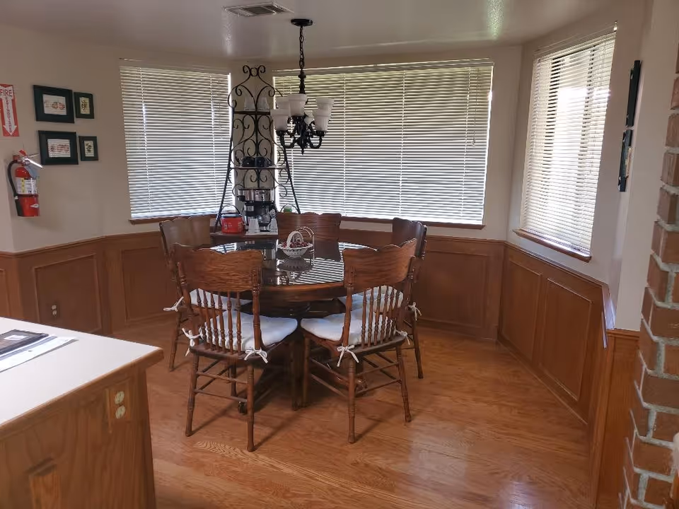 A dining area with a round wooden table surrounded by six wooden chairs with white cushions. The room has wood paneling on the walls, three windows with closed blinds, a chandelier hanging above the table, and a metal shelving unit with a coffee maker and other items. A fire extinguisher is mounted on the wall to the left.