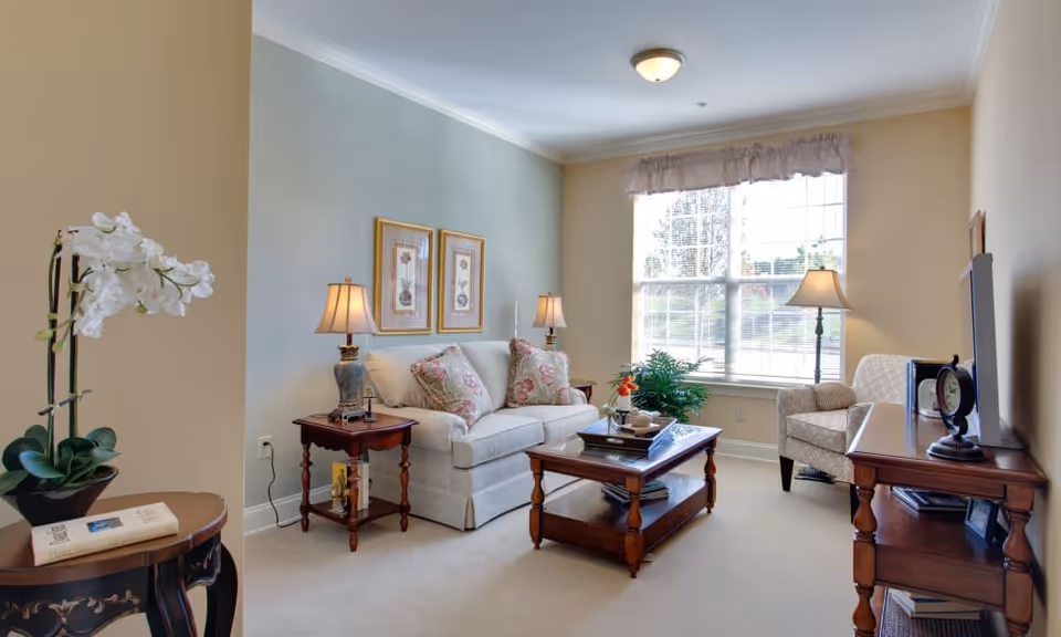 A cozy living room with a white sofa adorned with floral cushions, two wooden side tables with lamps, a wooden coffee table with decorative items, a patterned armchair near a large window with sheer curtains, and a wooden TV stand with a television and clock. A potted orchid is on a small round table in the foreground.