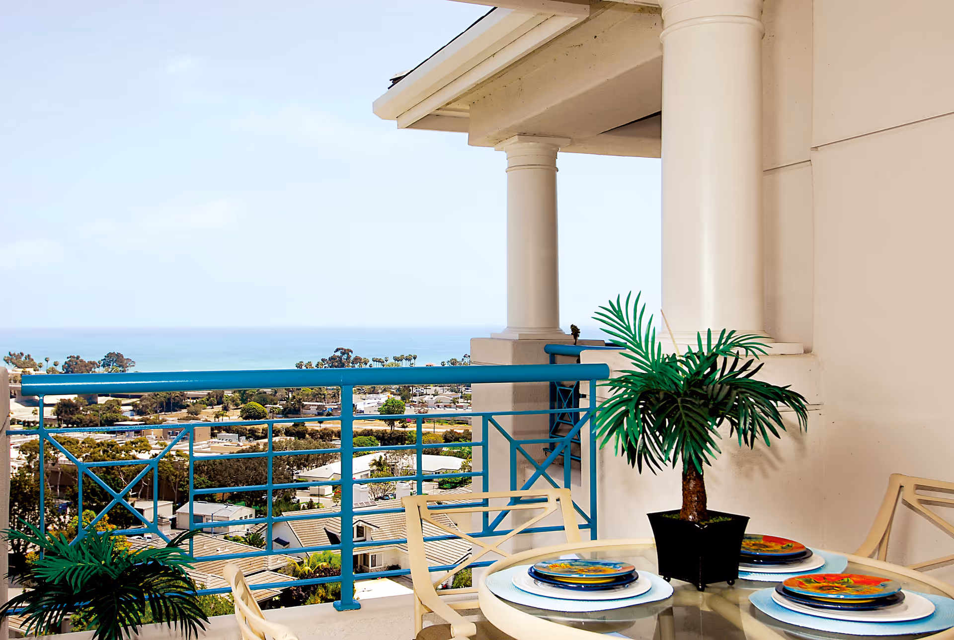 Balcony with a round glass table set with colorful plates and a potted plant, overlooking a coastal town with the ocean in the background under a clear sky.