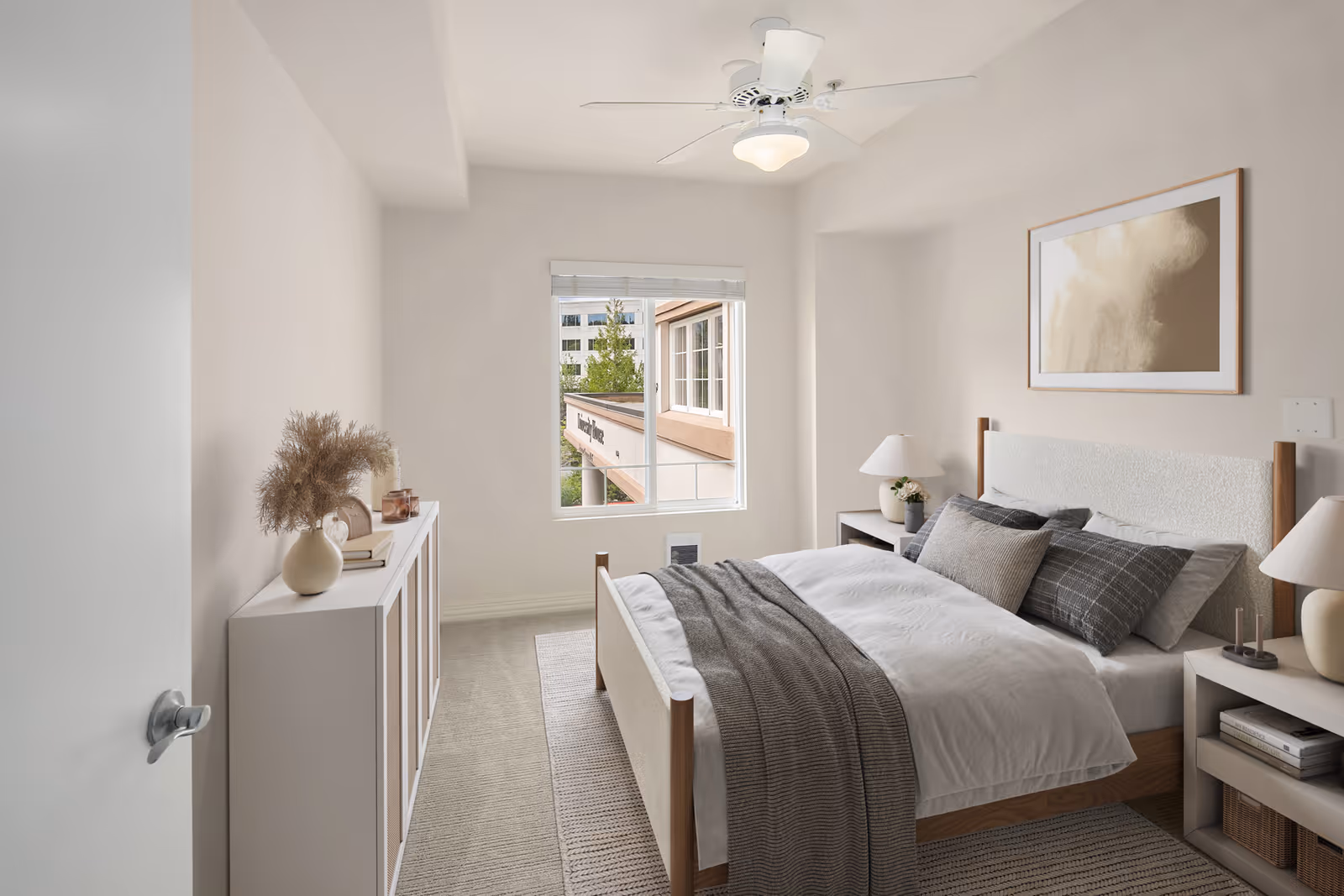 A bright and cozy bedroom with a neatly made bed featuring white and gray bedding. The room has a ceiling fan with a light, a window showing an exterior view, two bedside tables with lamps, a framed abstract artwork on the wall, and a white cabinet with decorative items including a vase with dried plants.