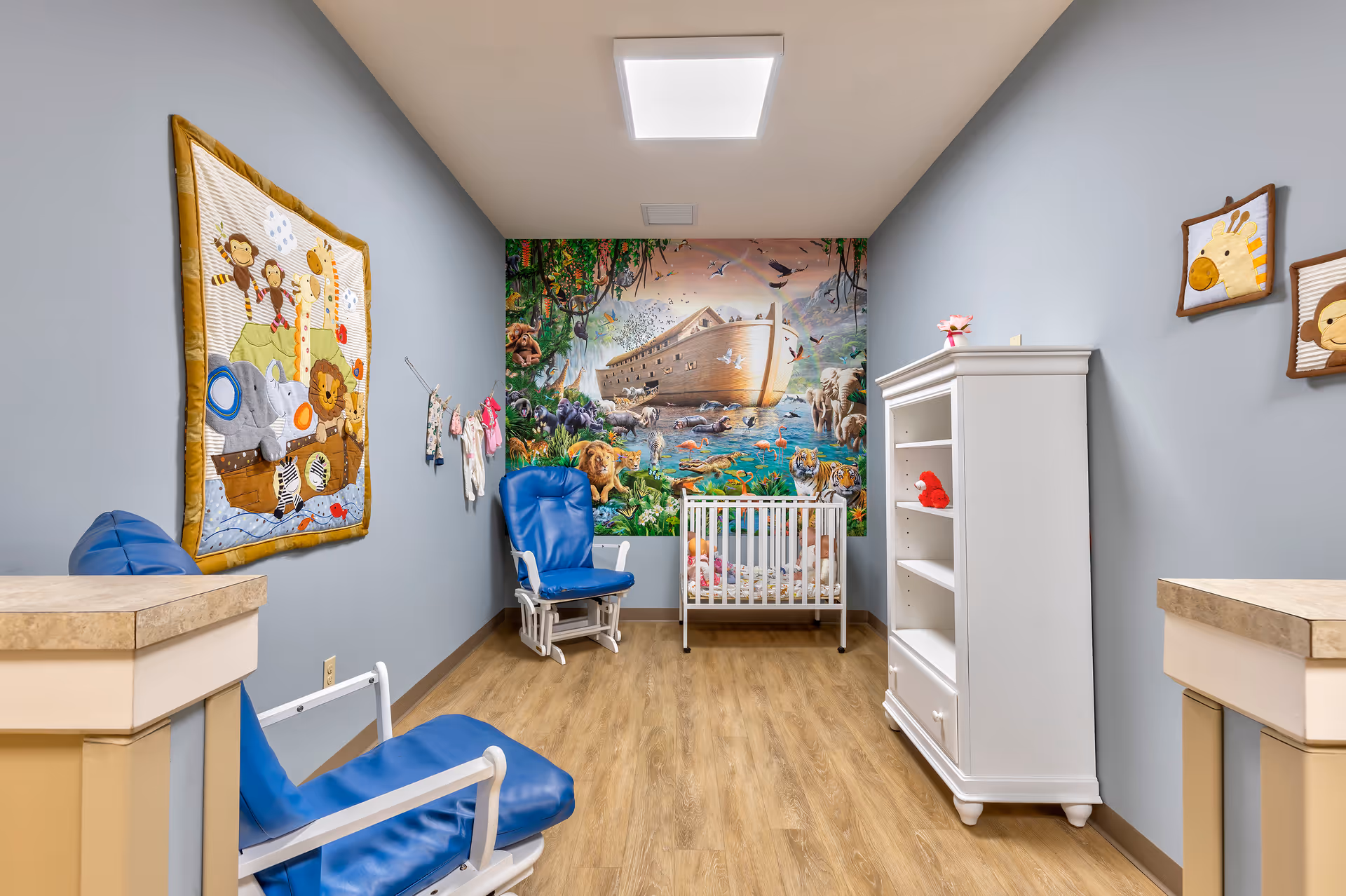 A brightly decorated nursery with blue rocking chairs, a white crib and bookshelf, and a colorful Noah's Ark mural on the back wall.