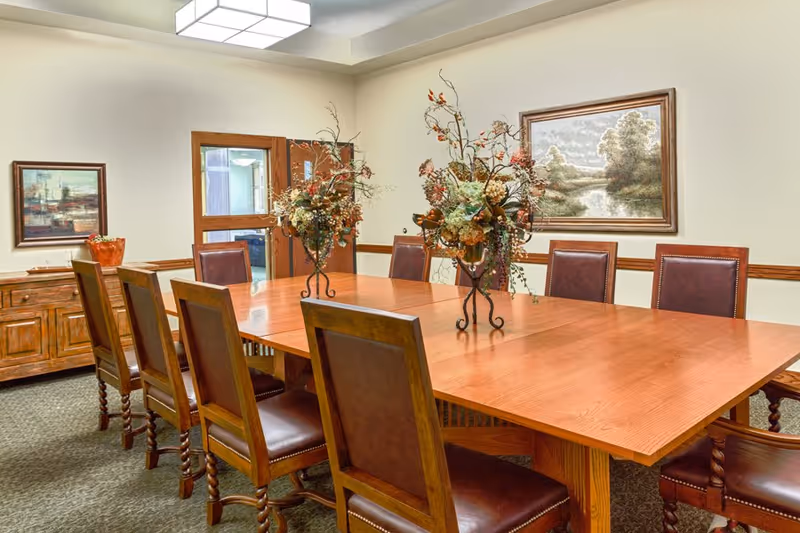 Long wooden table surrounded by leather-upholstered chairs with floral centerpieces in a well-lit dining/meeting room with framed landscape paintings.