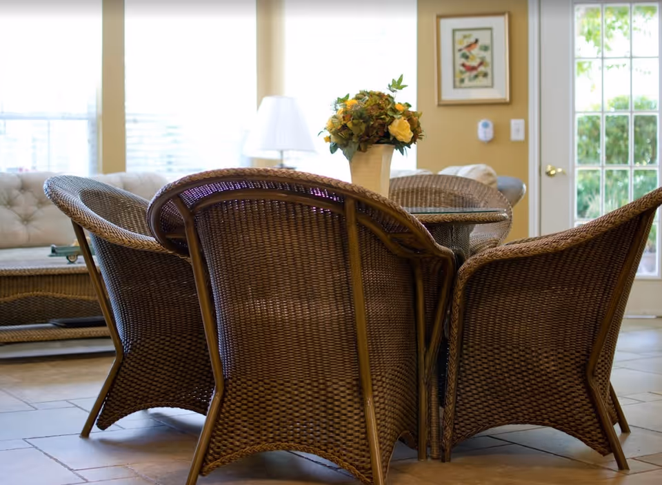 Wicker dining table surrounded by woven chairs and a vase of flowers in a bright room with windows and a door.