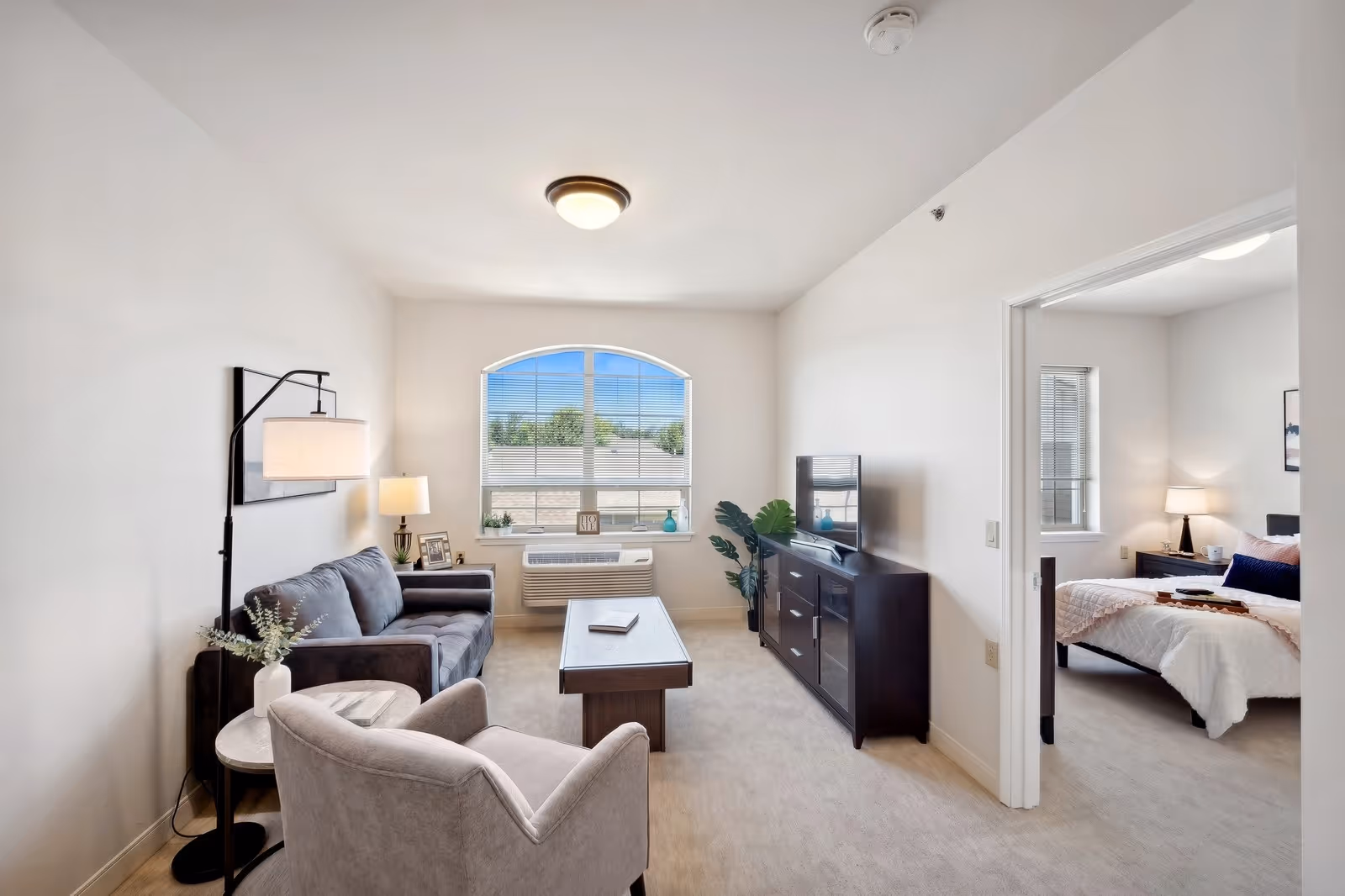 A bright living room with a gray sofa, a beige armchair, a wooden coffee table, and a TV on a dark wooden stand. There is a large window with blinds letting in natural light, and a floor lamp and table lamp providing additional lighting. To the right, an open doorway leads to a bedroom with a bed, nightstand, and lamp.