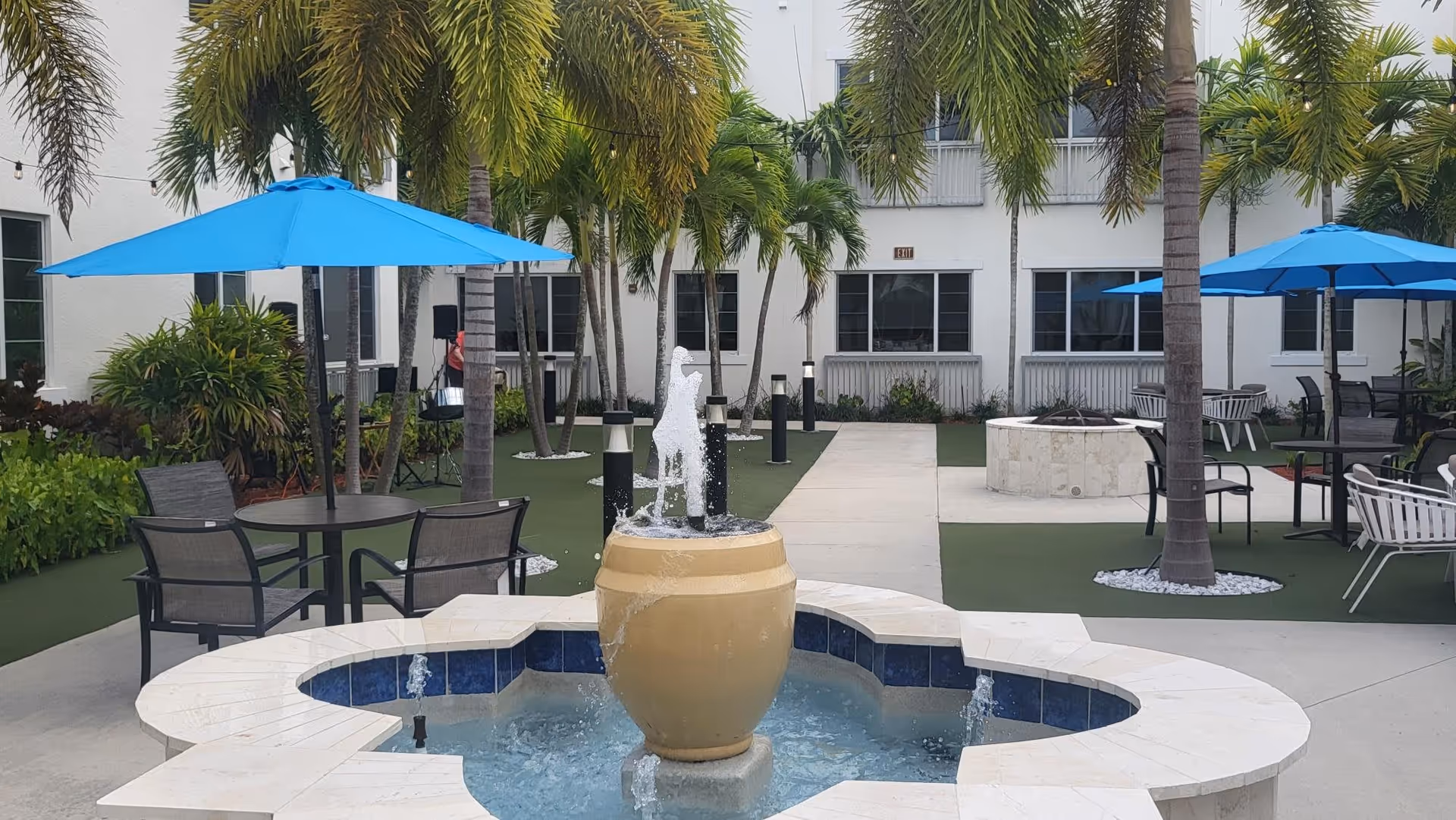 Outdoor courtyard area with a central water fountain featuring a large beige urn with water spouting from the top and sides. Surrounding the fountain are palm trees, green artificial turf, and several seating areas with tables, chairs, and blue umbrellas. The background shows a white building with multiple windows and an exit sign above a door.