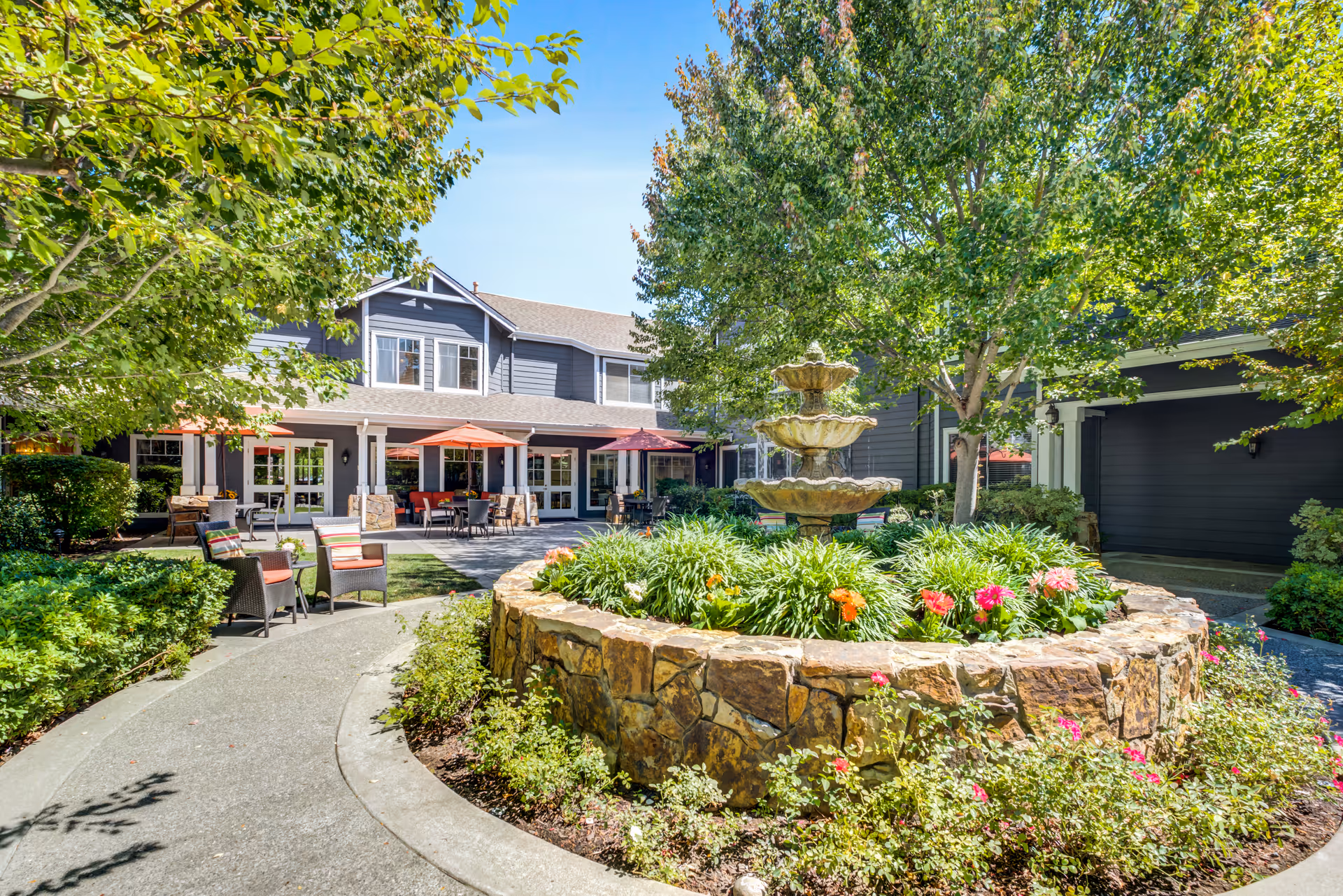 Outdoor courtyard area at Aegis Living Moraga featuring a multi-tiered stone fountain surrounded by greenery and colorful flowers. There are trees providing shade, a curved concrete pathway, and patio seating with tables and chairs under red umbrellas in front of a two-story building with gray siding and white trim.