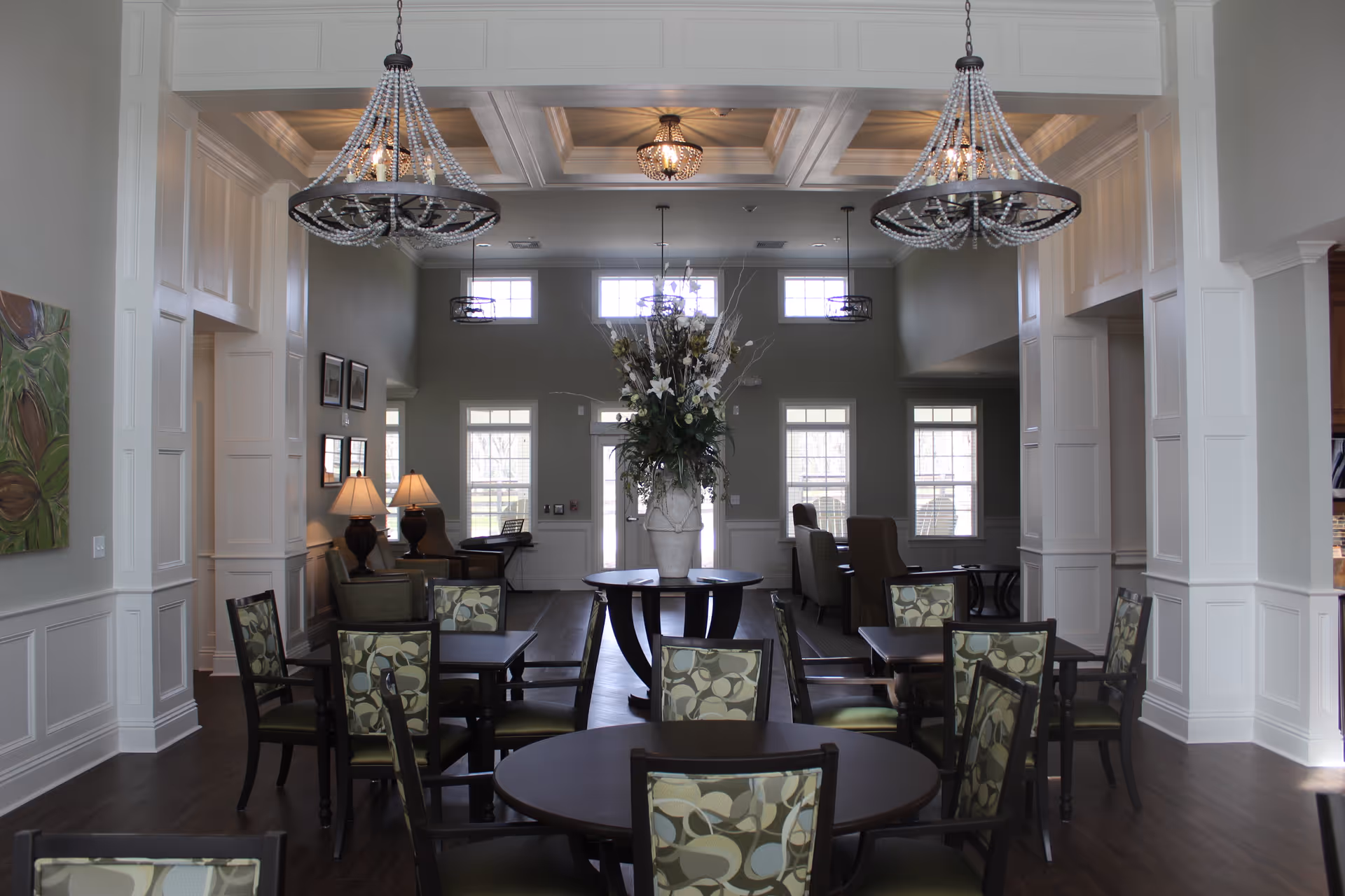 Bright, elegant communal dining room with round tables and patterned chairs, chandeliers, and a central floral arrangement.