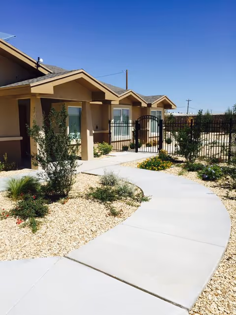 Curved concrete walkway leading to a gated entrance of a beige stucco building with small bushes and desert landscaping under a clear blue sky.