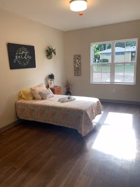 A simple bedroom with a single bed covered in a floral bedspread, a yellow pillow and two decorative pillows. There is a small wooden nightstand with a plant and a decorative item. The walls are light-colored with a framed sign that says 'gather', a hanging plant, and a floral wall decoration. Sunlight streams in through a large window, illuminating the wooden floor.