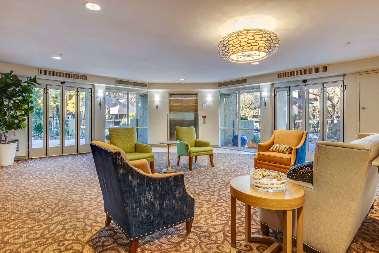 A spacious and well-lit common area with patterned carpet flooring, several upholstered chairs in green, orange, and blue, a small round table, and large windows and glass doors providing a view of an outdoor garden area. The ceiling features a decorative light fixture, and there is an elevator in the background.