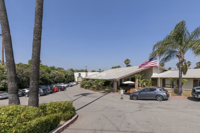 Front entrance of a single-story senior living facility with a covered drive, parked cars, palm trees, and an American flag.