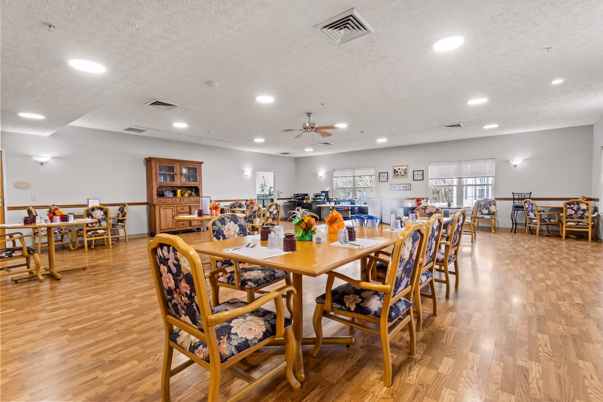 A spacious dining room with wooden floors and several wooden tables surrounded by floral upholstered chairs. The tables are set with placemats, utensils, glasses, and small floral centerpieces. The room has white walls, ceiling lights, a ceiling fan, and windows letting in natural light. A wooden cabinet is visible against one wall.