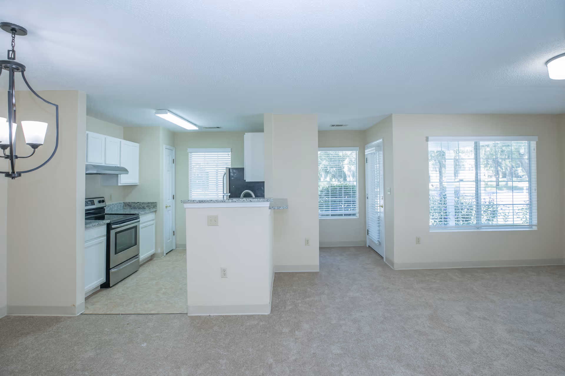 Interior view of a senior living facility showing a kitchen area with white cabinets, stainless steel stove, and granite countertops on the left. Adjacent to the kitchen is an open living space with beige carpet, large windows with blinds, and a door leading outside. The walls are painted light beige and the ceiling has a textured finish with ceiling lights.