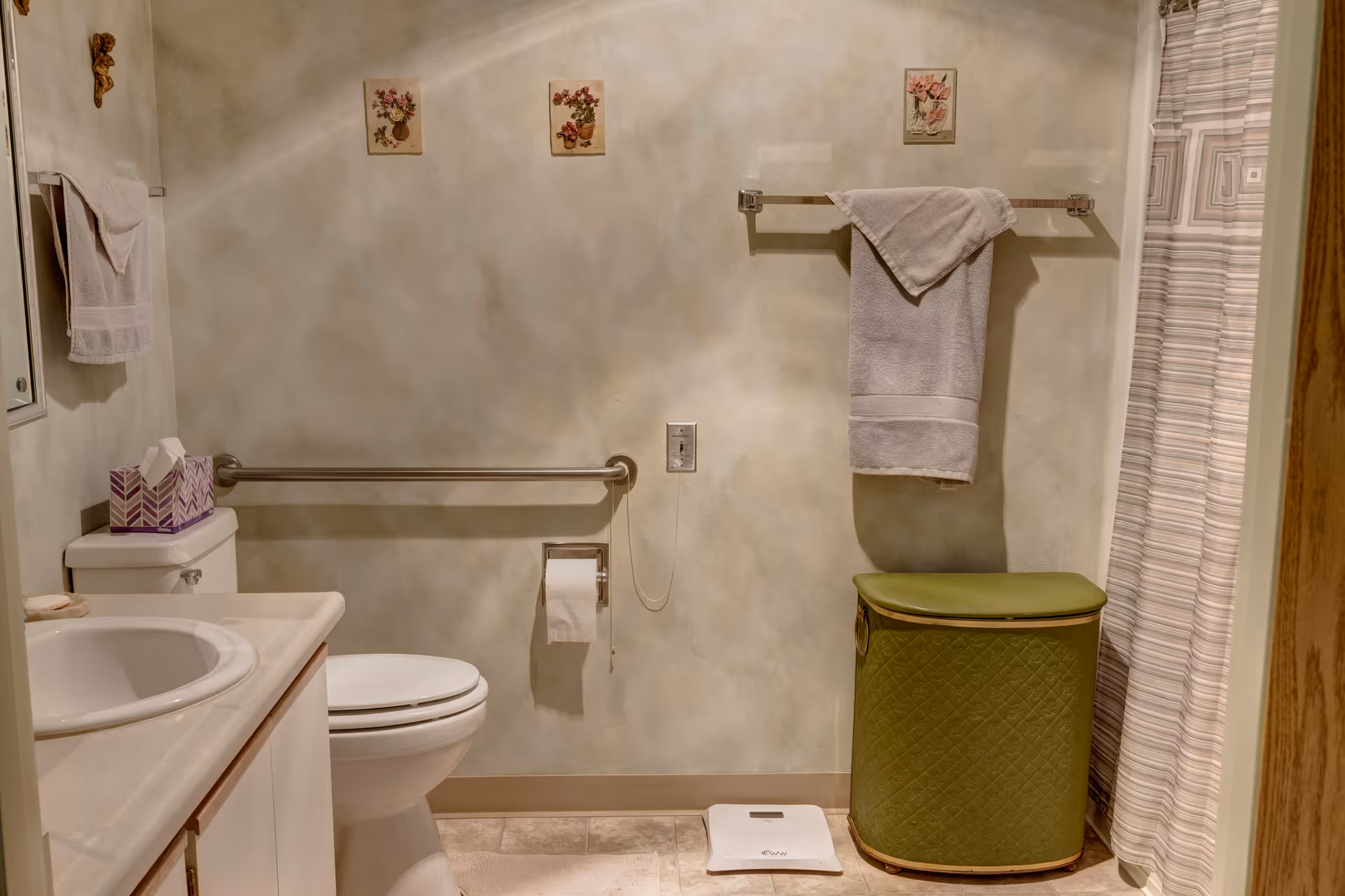Small accessible bathroom with a toilet, sink, grab bar, towel rack, laundry hamper, and shower curtain.