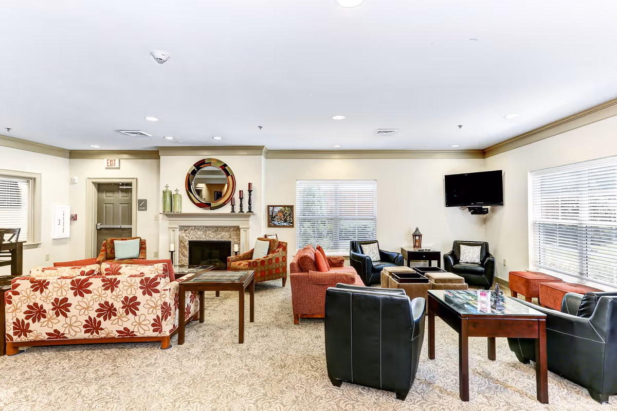 A bright and spacious living room area in an active adult community featuring multiple seating options including floral patterned sofas, red and black armchairs, a fireplace with a decorative round mirror above it, a wall-mounted TV, and large windows with blinds allowing natural light to fill the room.