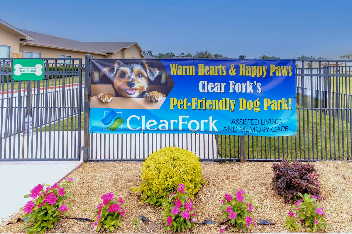 A fenced pet-friendly dog park at Clear Fork Assisted Living and Memory Care, featuring a banner with a happy dog and the text 'Warm Hearts & Happy Paws Clear Fork's Pet-Friendly Dog Park!'. There are colorful flowers and shrubs planted in front of the fence under a clear blue sky.