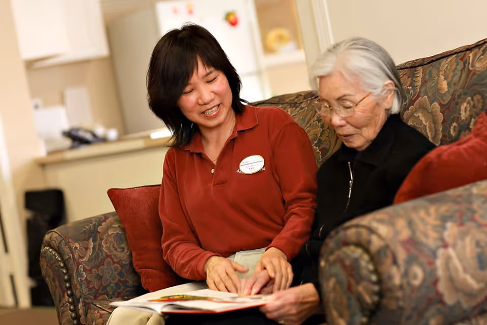 A caregiver in a red shirt is sitting on a patterned couch next to an elderly woman with white hair and glasses. They are looking at a book together and smiling in a cozy living room setting.