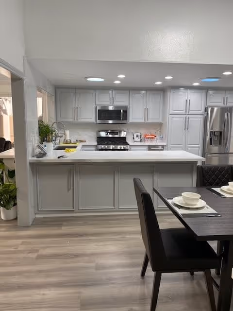 Modern kitchen with light gray cabinets, a stainless steel refrigerator, microwave, and stove. A white countertop island is in the center with a dining table and chairs nearby set with plates and bowls. The floor is light wood, and there are recessed ceiling lights.