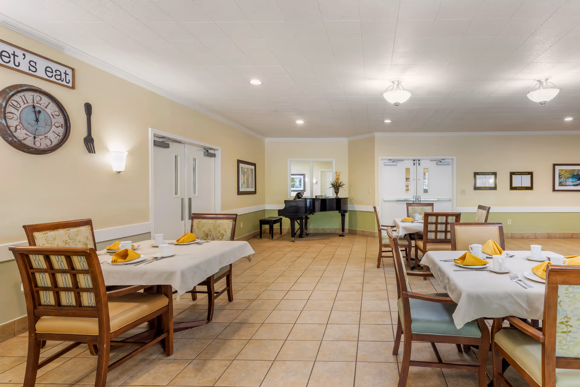 A dining room in a senior living facility with several tables set with white tablecloths, yellow napkins, cups, and utensils. The room has tiled floors, beige walls with a green lower section, and a ceiling with recessed lighting and two ceiling light fixtures. A black grand piano with a stool and a flower arrangement is positioned against the far wall. On the left wall, there is a clock and a sign that says 'let's eat'.