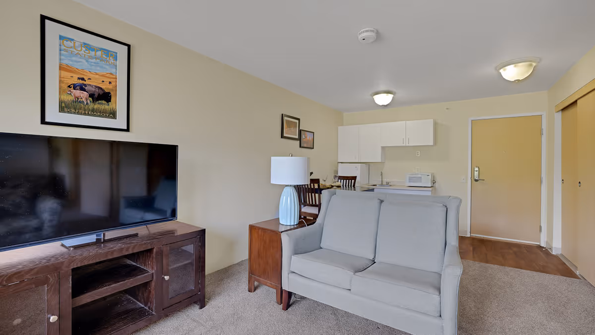 Interior view of a senior living facility room at Lodge At White Bear featuring a gray loveseat, a wooden TV stand with a flat-screen TV, a small dining table with chairs, a kitchenette with white cabinets, a microwave, and a refrigerator. The walls are decorated with framed pictures, and the floor is carpeted with a section of wood flooring near the entrance door.