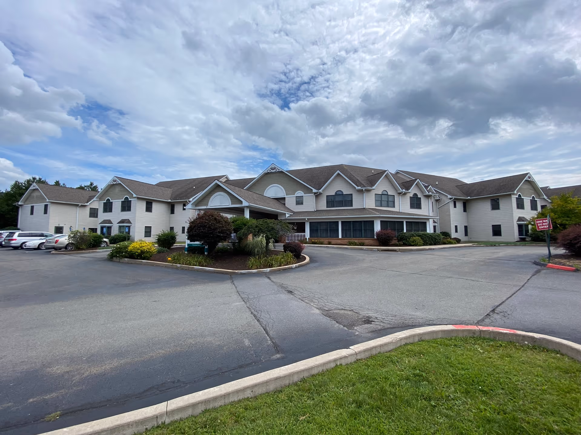Front exterior of a two-story senior living facility with a circular driveway, landscaping, parked cars and a cloudy sky.