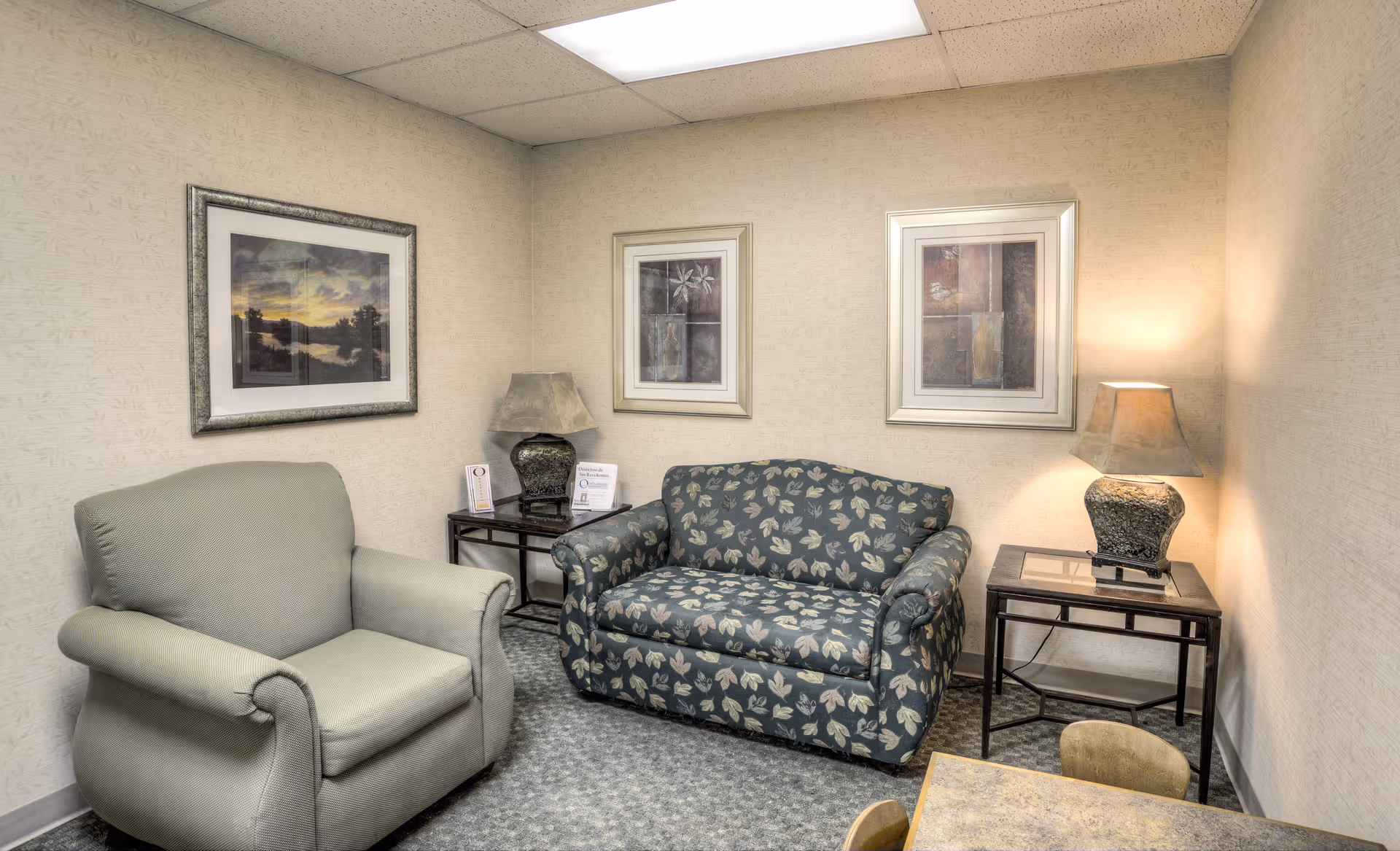 A small sitting area with a green upholstered armchair and a floral patterned loveseat. There are two side tables with lamps on either side of the loveseat. Three framed pictures hang on the beige walls. The floor is carpeted and there is a small table with chairs partially visible in the foreground.