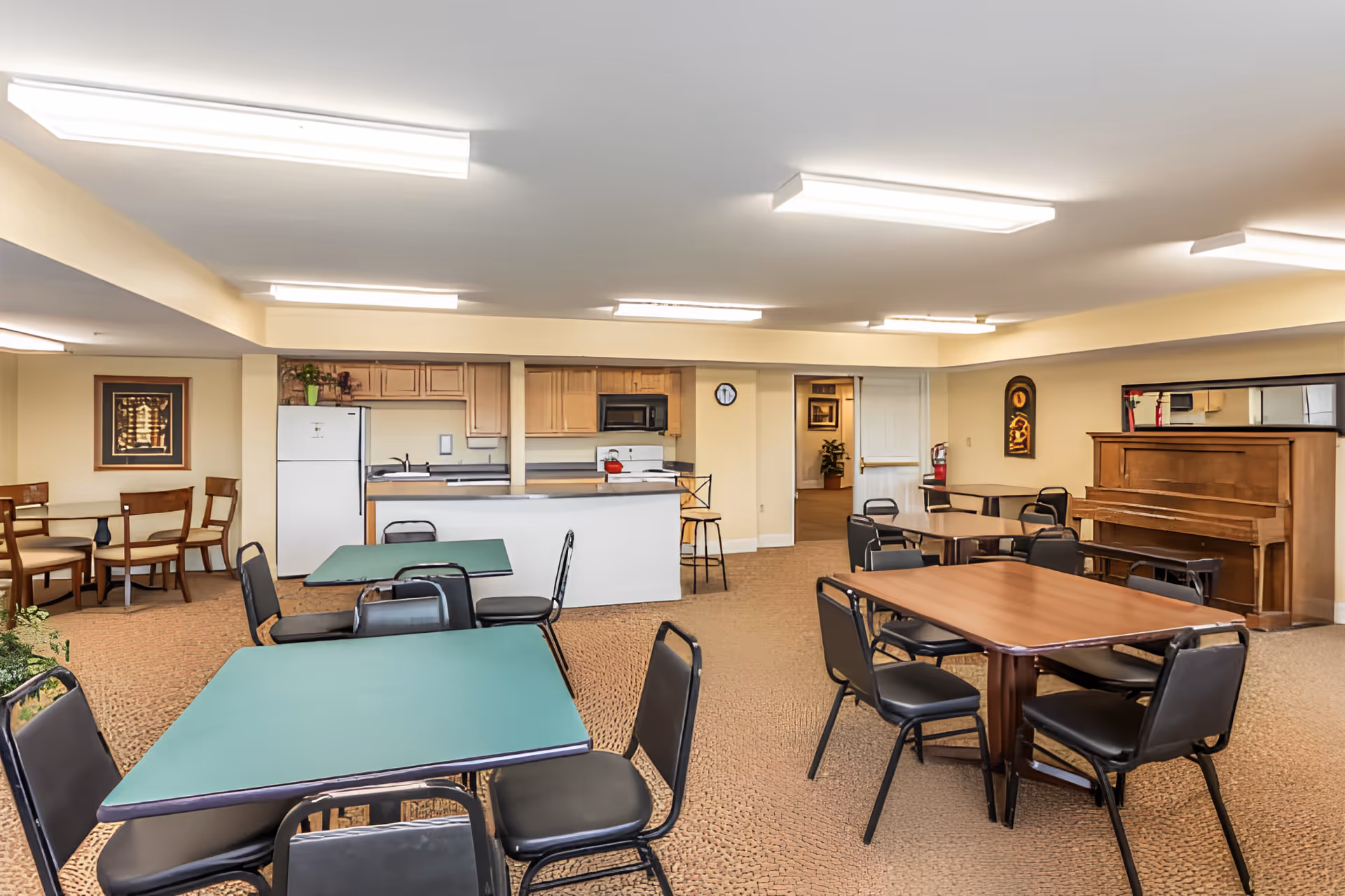 Bright communal dining/activity room with multiple tables and chairs, a kitchenette and a piano.