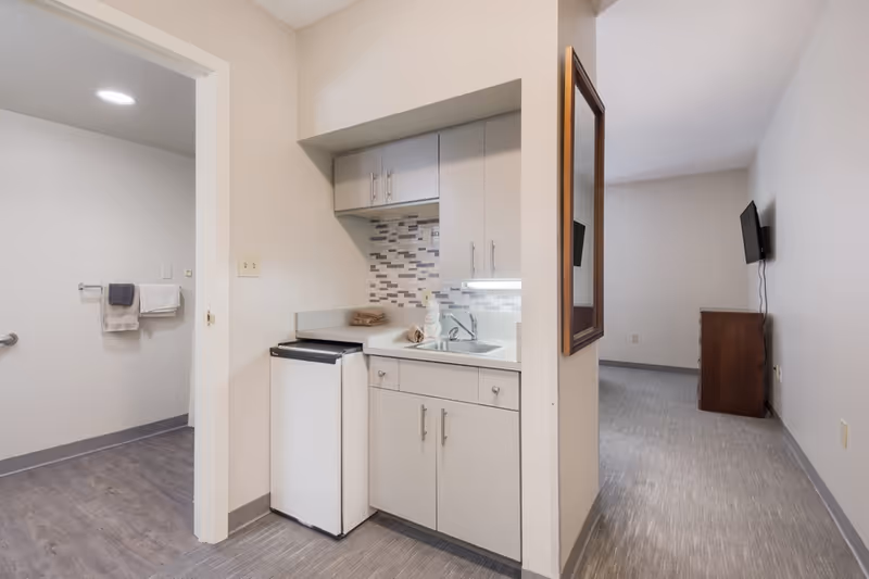 Interior view of a senior living facility room showing a small kitchenette with white cabinets, a mini refrigerator, a sink, and a tiled backsplash. To the left is an open doorway leading to a bathroom with towels hanging on a rack. To the right is a hallway with a mounted flat-screen TV and a wooden dresser against the wall.