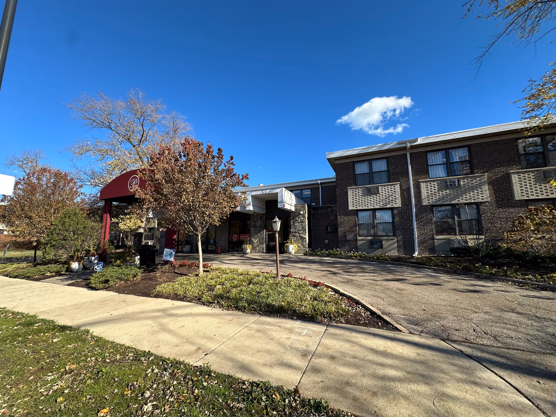 Front entrance of a two-story brick health care building with a maroon awning, driveway, and landscaped trees under a clear blue sky.