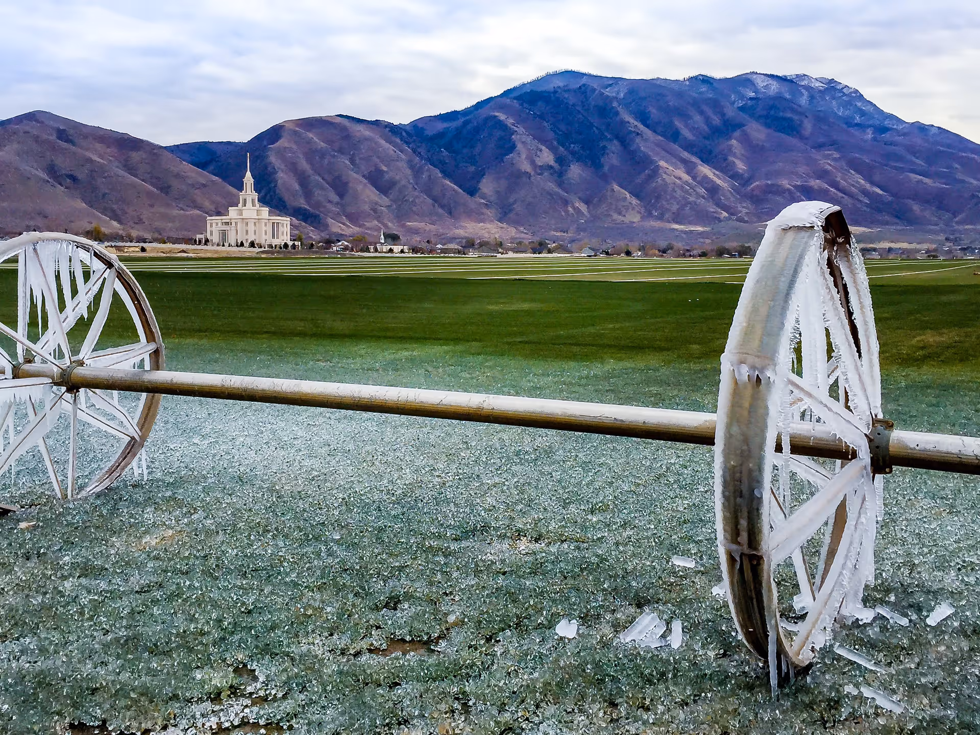 Icy irrigation wheel on a frosted field with a distant white building and mountains under a cloudy sky.