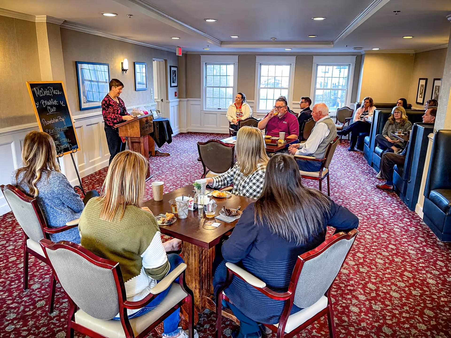 Group of people seated around tables in a carpeted dining room listening to a woman speaking at a podium.