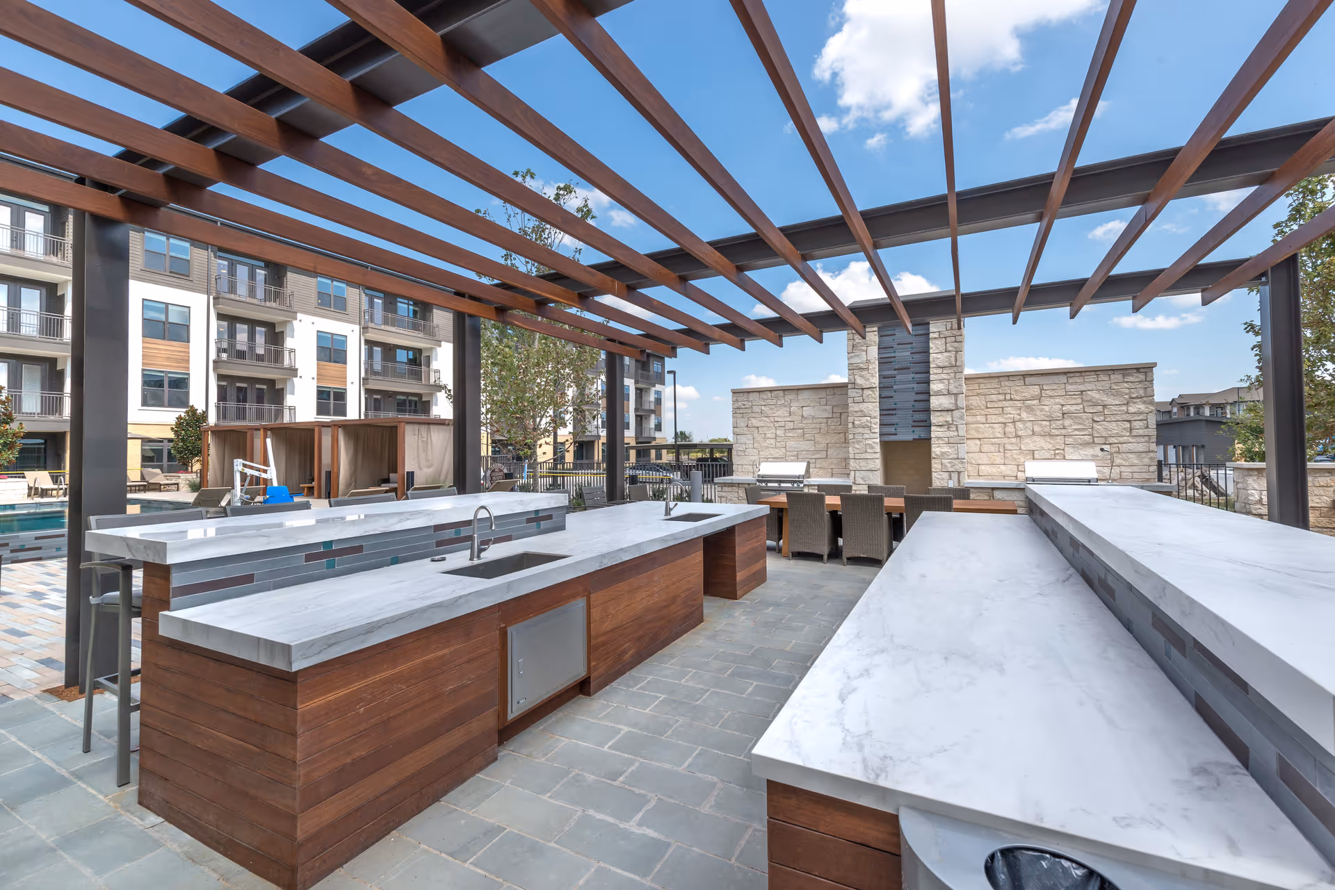 Outdoor kitchen area with marble countertops, sinks, and seating under a wooden pergola. In the background, there are lounge chairs, cabanas, and a stone wall with grills. Apartment buildings and a blue sky with clouds are visible.