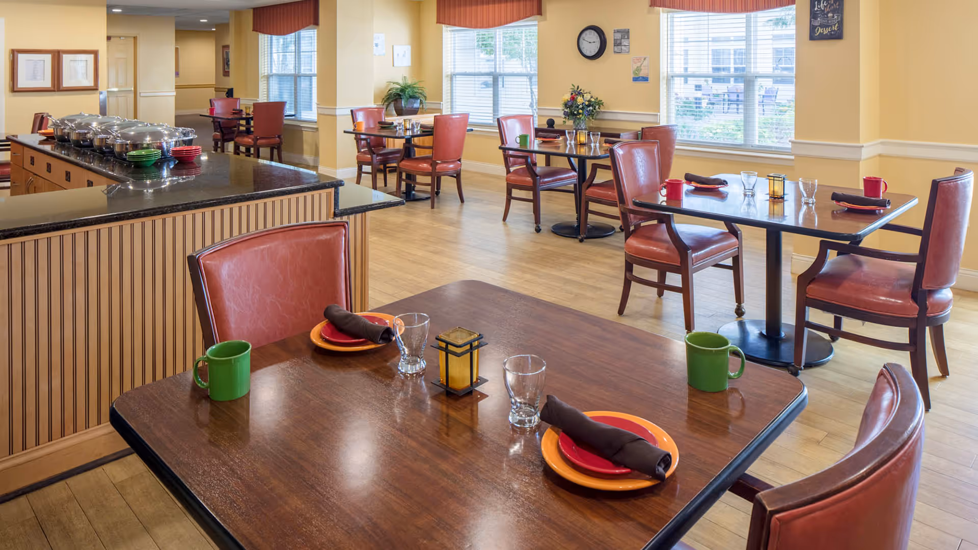 A dining area in a senior living facility with several tables set with plates, cups, glasses, and napkins. The room has wooden floors, large windows with red valances, and red cushioned chairs. A buffet counter with covered dishes is visible on the left side of the image.