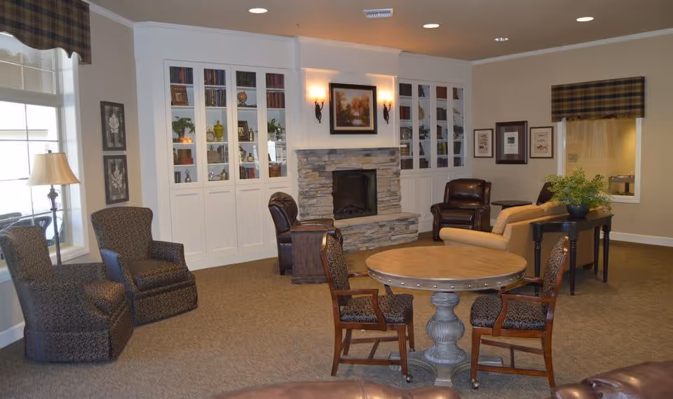 A cozy living room area in an assisted living facility featuring a stone fireplace with a framed painting above it, built-in white cabinets with glass doors displaying books and decorative items, several upholstered armchairs, a round wooden table with four chairs, a sofa, and a side table with a potted plant. The room has beige walls, carpeted floor, and windows with plaid valances.