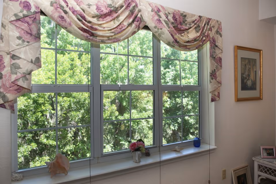 A window with white frames and floral valance curtains in shades of pink and purple. Outside the window, green leafy trees are visible. The windowsill holds a large seashell, a small vase with pink flowers, a starfish, and a blue glass candle holder. On the right wall, there is a framed painting and a small table with a framed photo and other items.